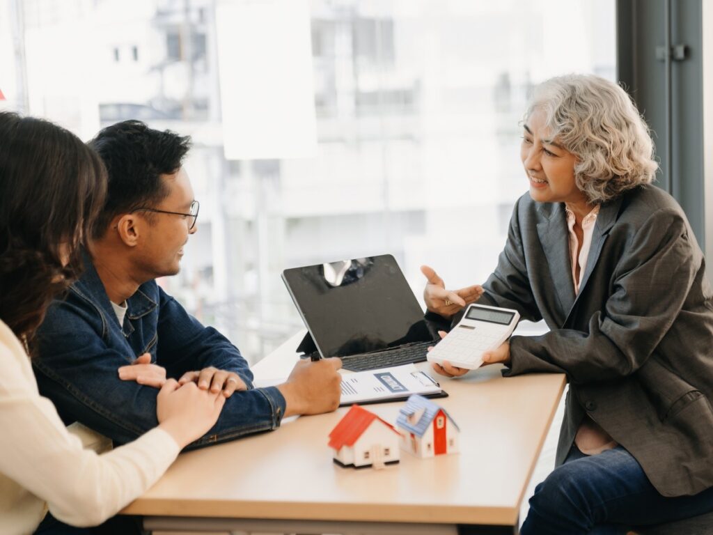 Meeting with an Agent About a Home Purchase Text: Real estate agent with a calculator meeting a young couple at a desk with a contract and model houses
