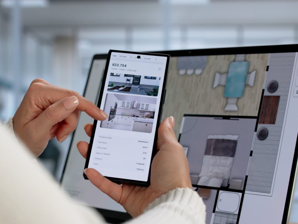 Browsing Property Listings on a Phone Person browsing a real estate listing on a smartphone with a floor plan shown on a monitor behind