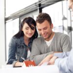 Smiling young couple signing paperwork with a real estate agent, a model house on the desk